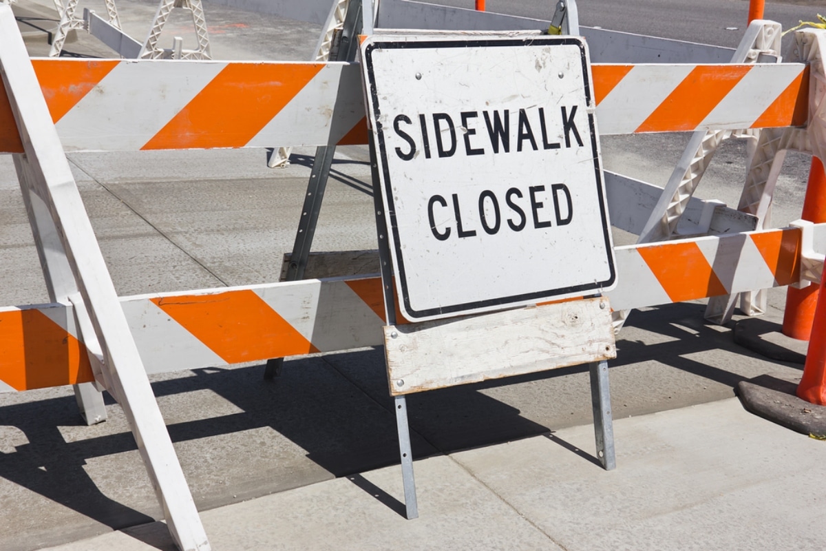 A barrier with sign stating 'Sidewalk Closed' to direct pedestrians around construction site.