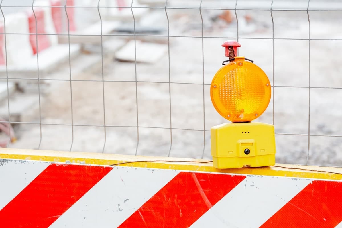 Red and white barricade, with yellow light outside fenced construction site.