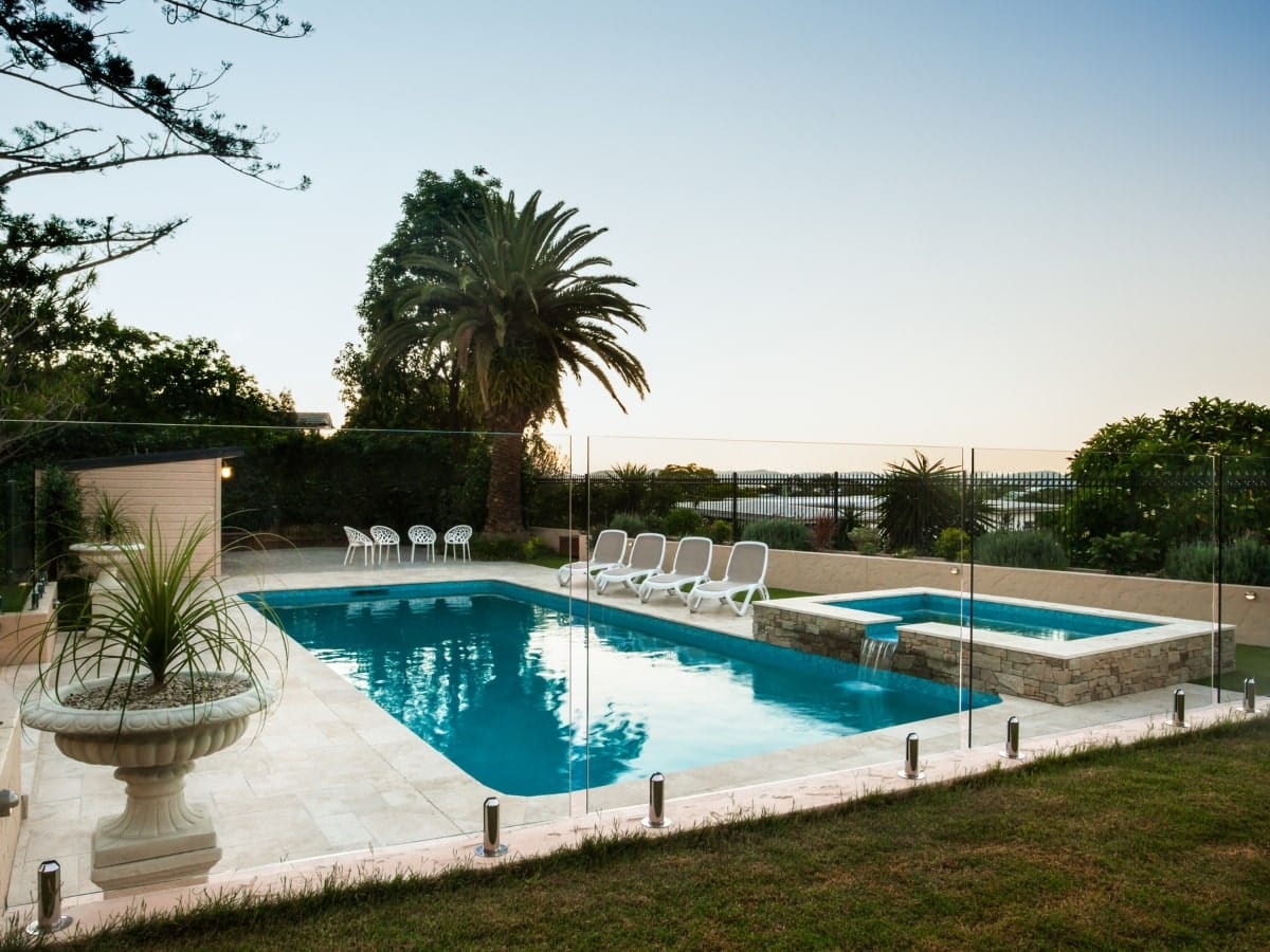 Elegant swimming pool encircled by clear glass fencing, showcasing one of the stylish types of glass pool fencing set amidst lush greenery and comfortable poolside loungers.