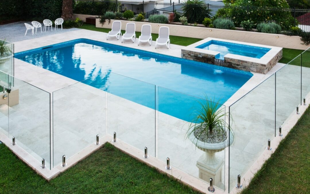 A modern swimming pool showcasing a glass panel fence, one of the popular types of pool fencing, surrounded by a green lawn, trees, and outdoor chairs.