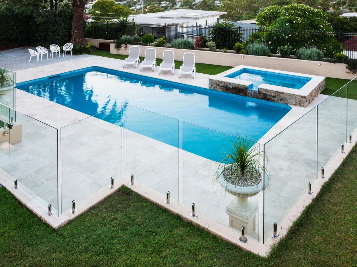 A modern swimming pool showcasing a glass panel fence, one of the popular types of pool fencing, surrounded by a green lawn, trees, and outdoor chairs.