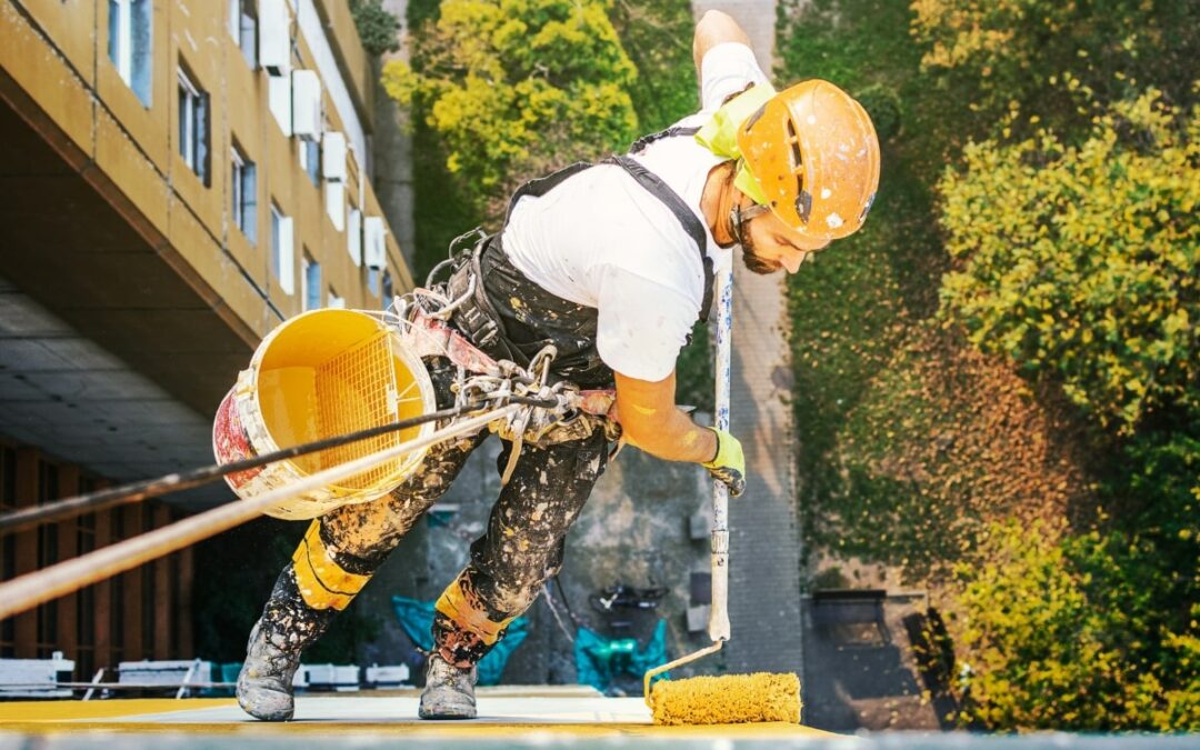 A rope access technician demonstrates working safely at heights, secured with harness and helmet, while applying paint on a building's exterior.